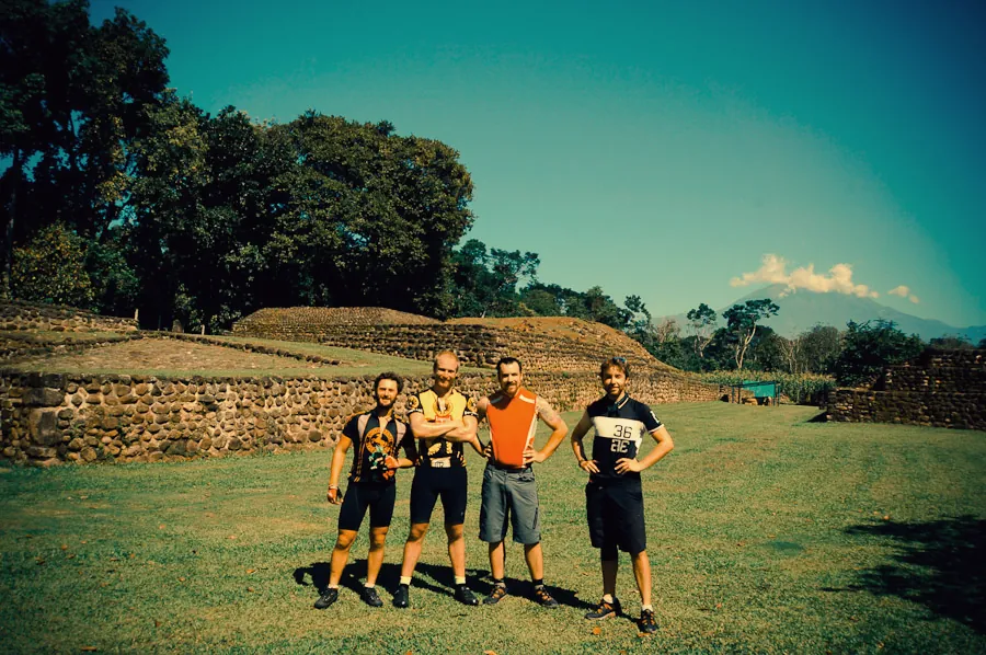 The four riders in front of ancient Maya mounds, with a volcano smoking behind them.