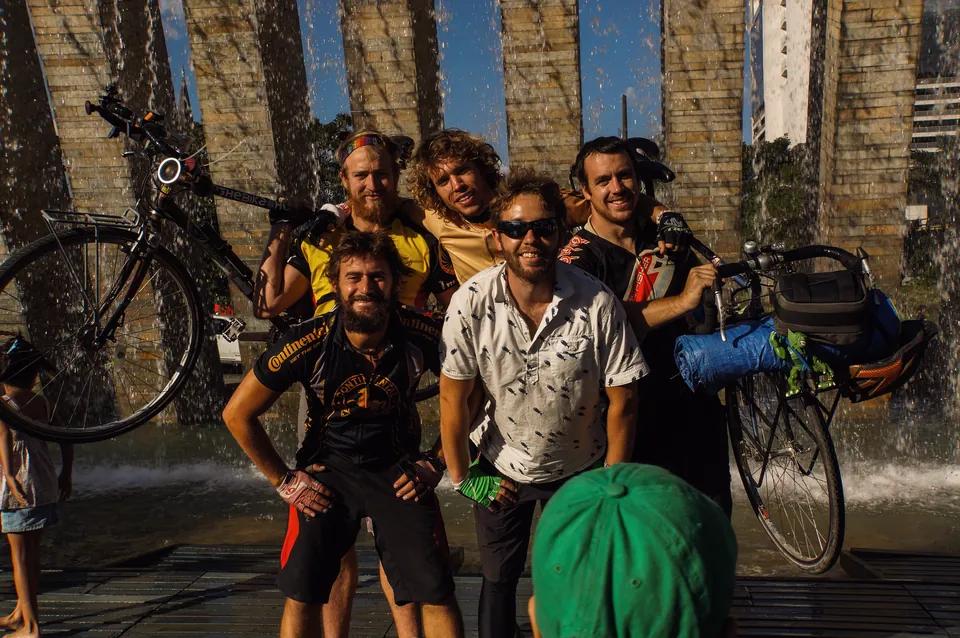 Five riders holding their loaded bicycles above their heads by a stone fountain in Medellín.