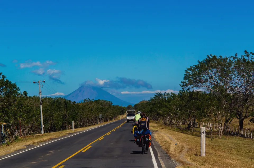 Two riders on a straight road heading towards a volcano under a deep blue sky in Nicaragua.