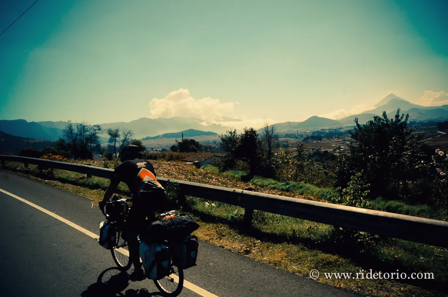 A rider on a guard-railed mountain road with volcanoes in the distance.