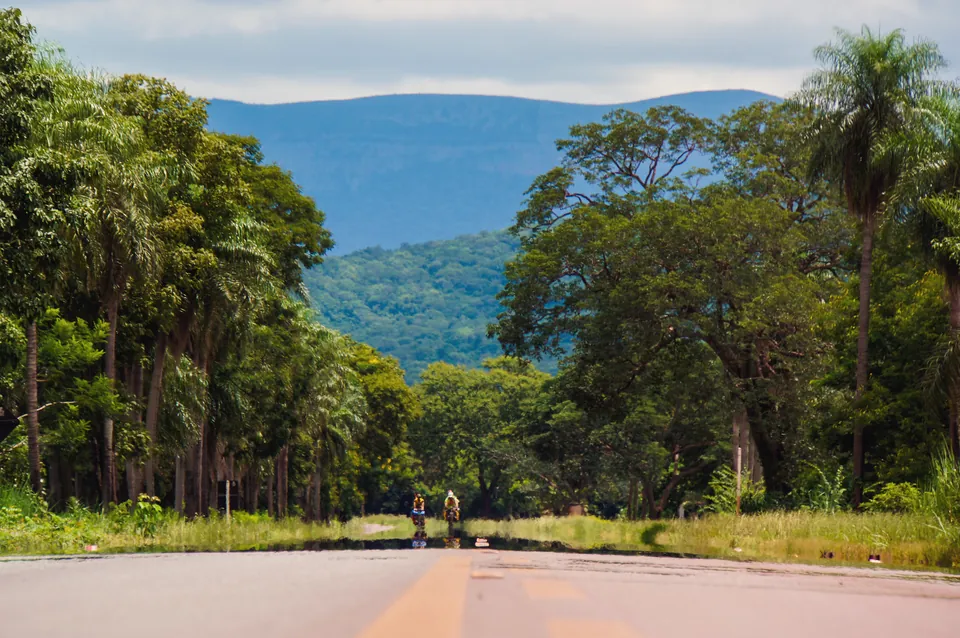 Two tiny riders on a palm-lined road below a blue escarpment in the heat.
