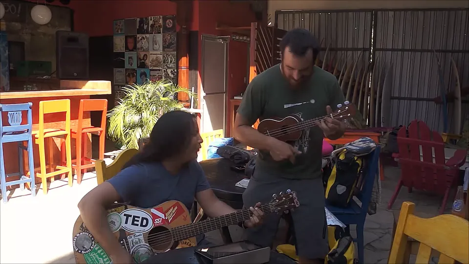 Two men playing guitar and ukulele together at a hostel bar.