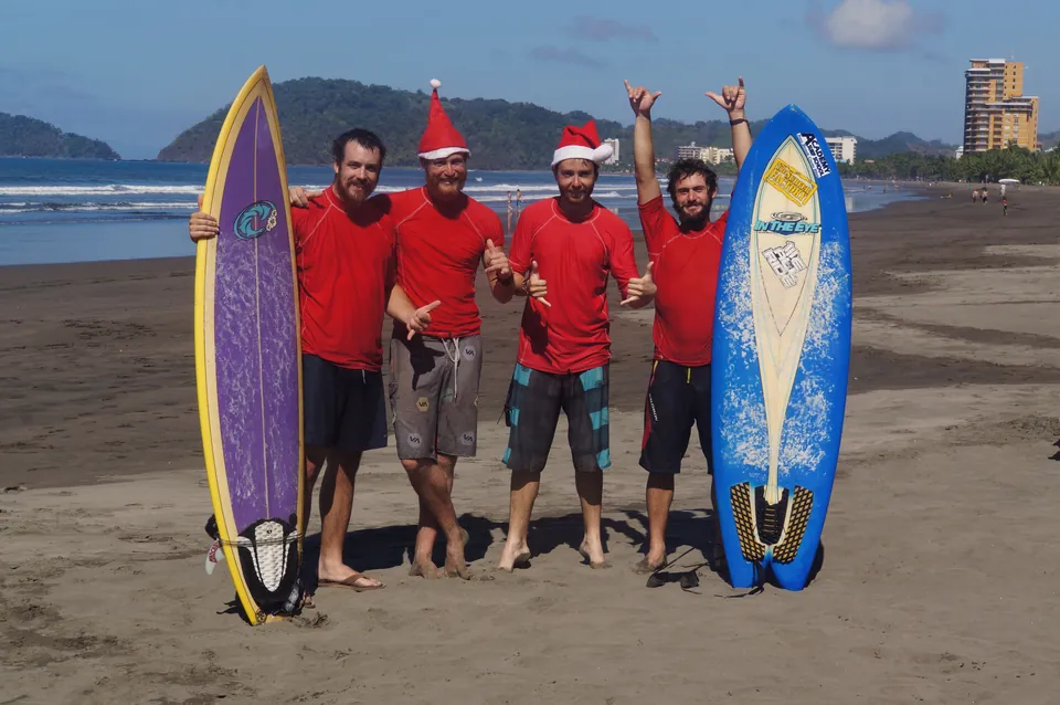 Four riders in red rash vests, two in Santa hats, holding surfboards on a dark-sand beach.