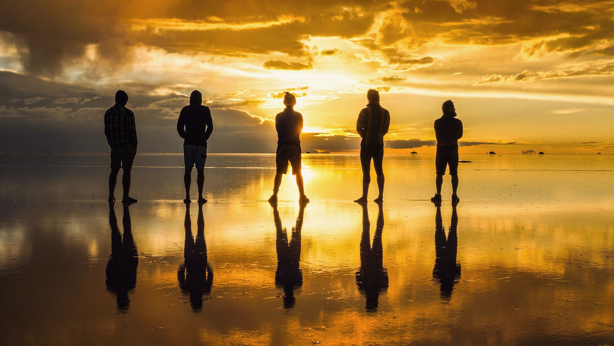 Five friends stand in silhouette against a golden sunset on the salt flats, reflected in the water.