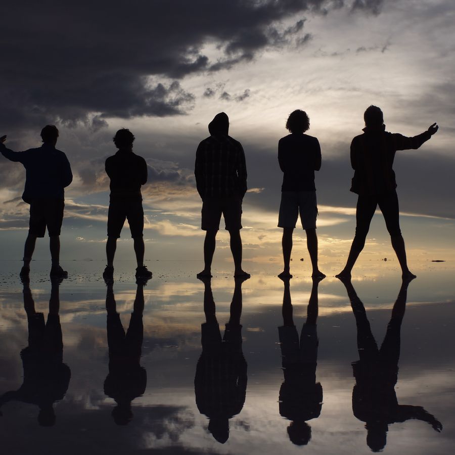 Five riders stand in silhouette on the flooded Salar de Uyuni at sunset, reflected in the water.