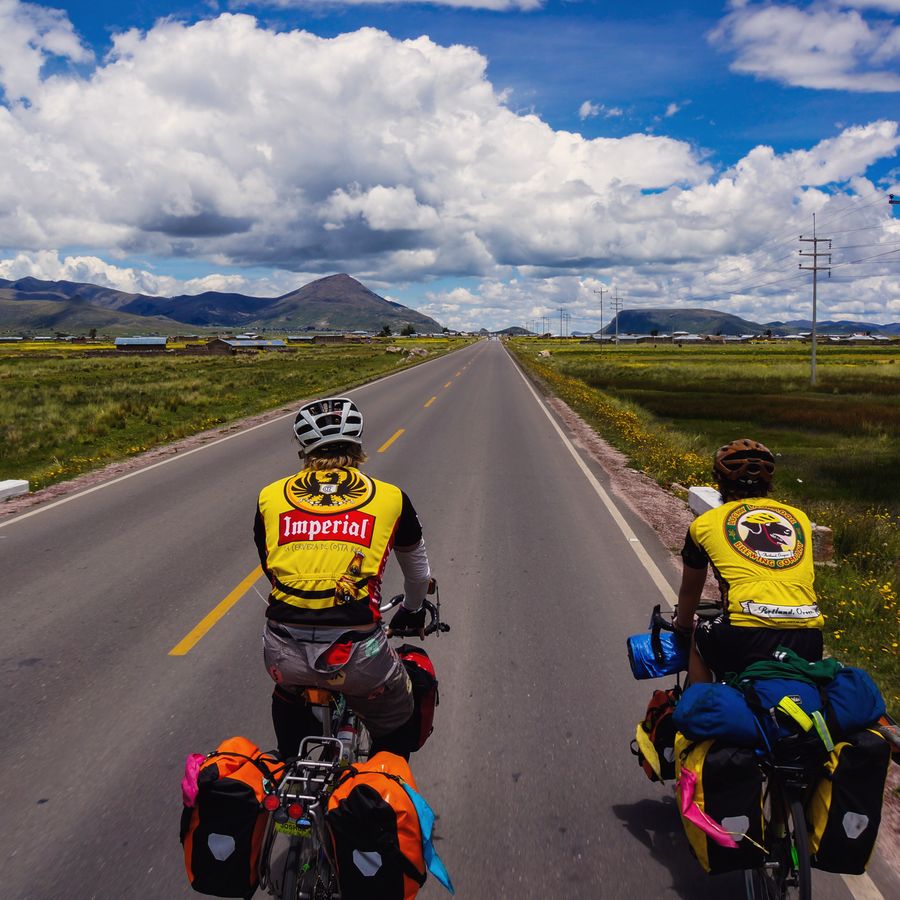 Two loaded touring bikes on a dead-straight road across the Andean altiplano under huge white clouds.